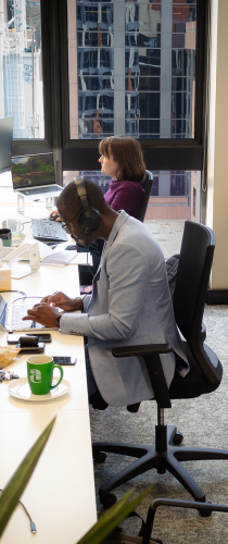 Team members working at their city office desks, with Allen + Clarke branded mugs and urban views through large windows providing natural light.