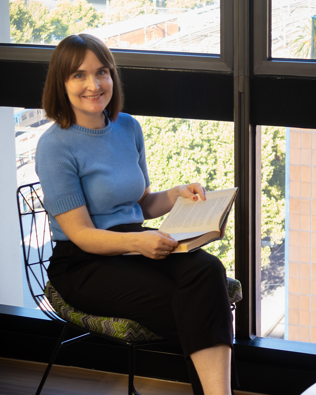 Woman in blue sweater holding open book by window, seated in patterned chair