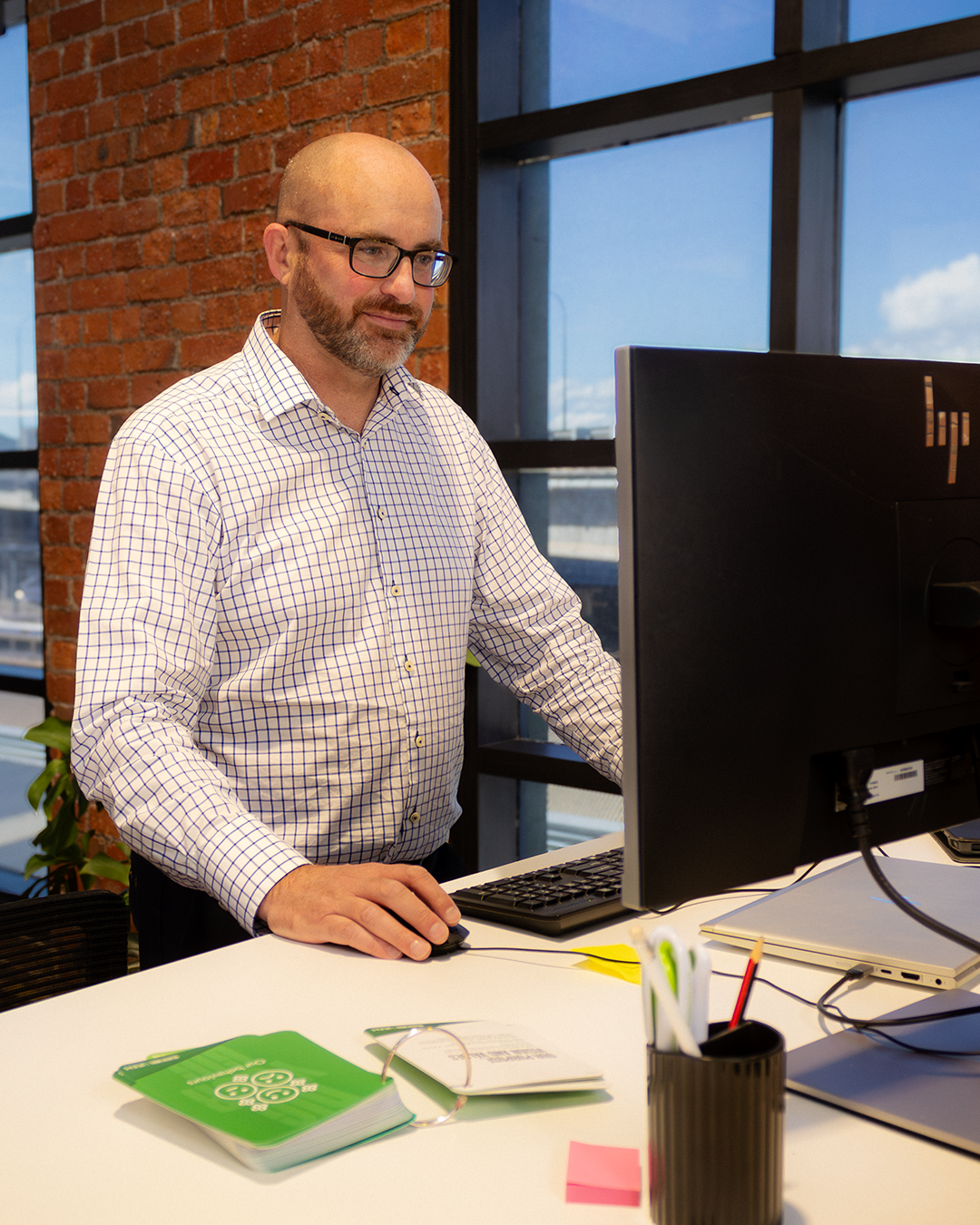 Professional in checkered shirt at standing desk with monitor, exposed brick wall and windows behind. 