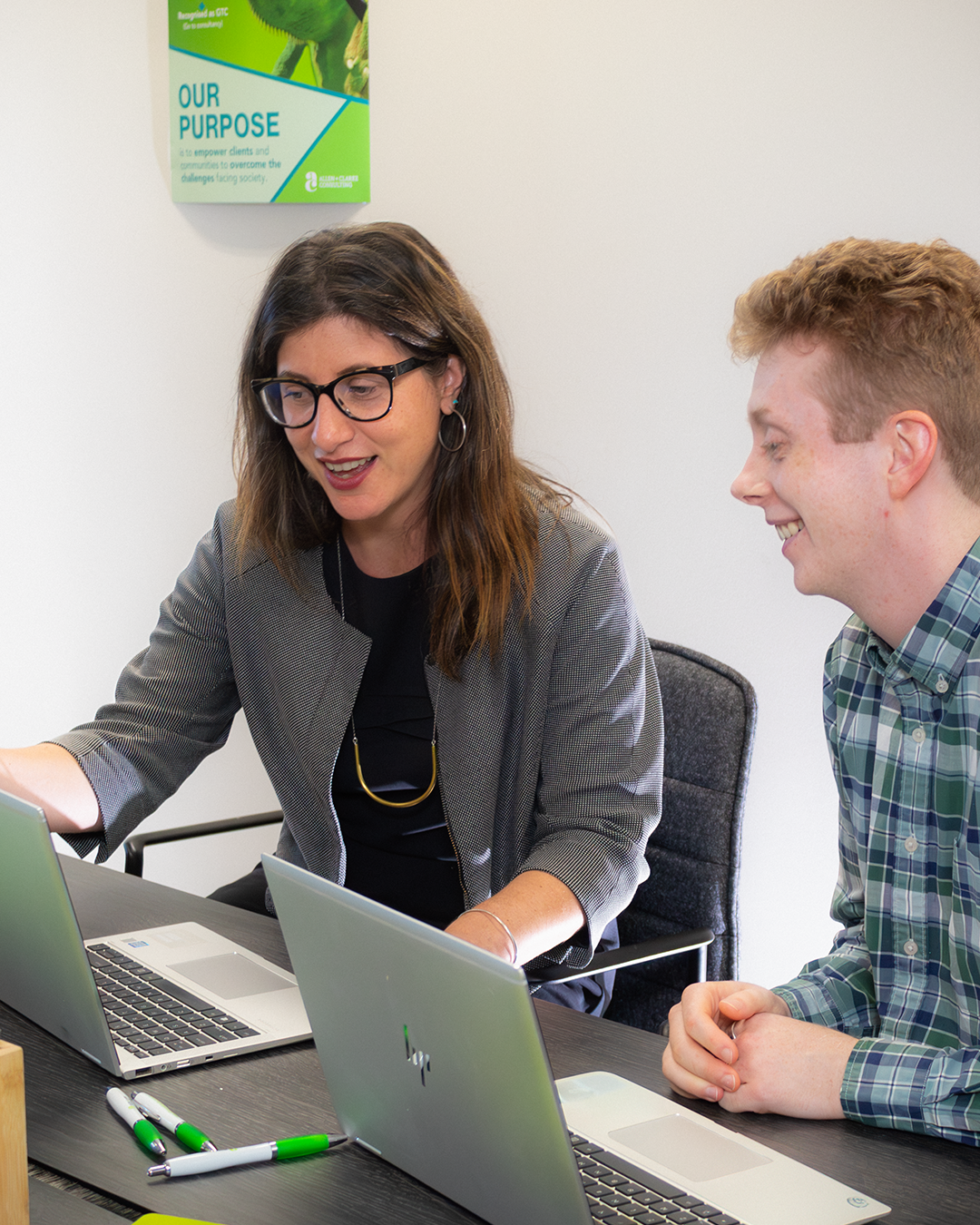 Two professionals smiling during collaboration at laptop, green "Our Purpose" poster visible on wall behind them
