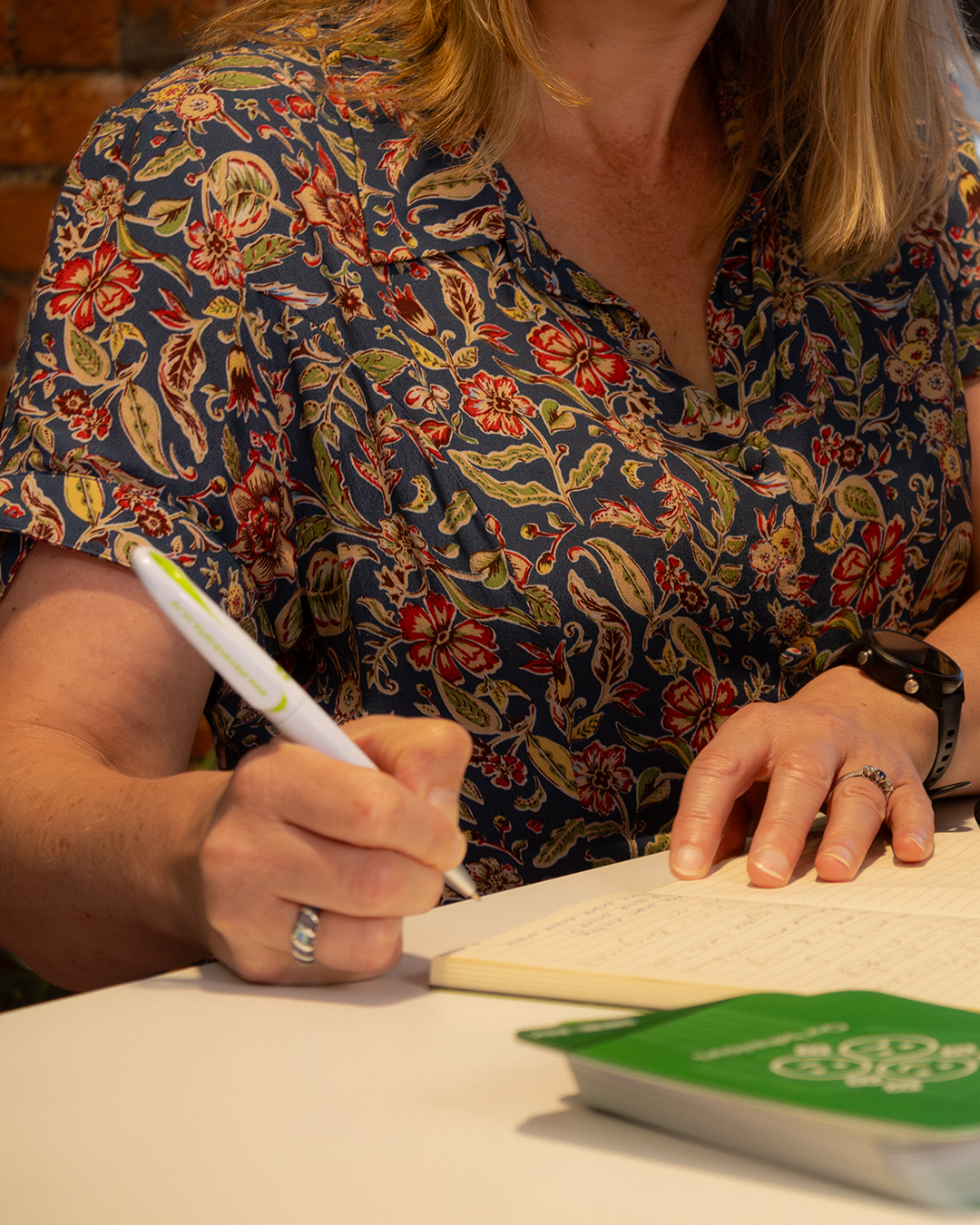 Woman in floral navy blouse writing in notebook, wearing watch and ring