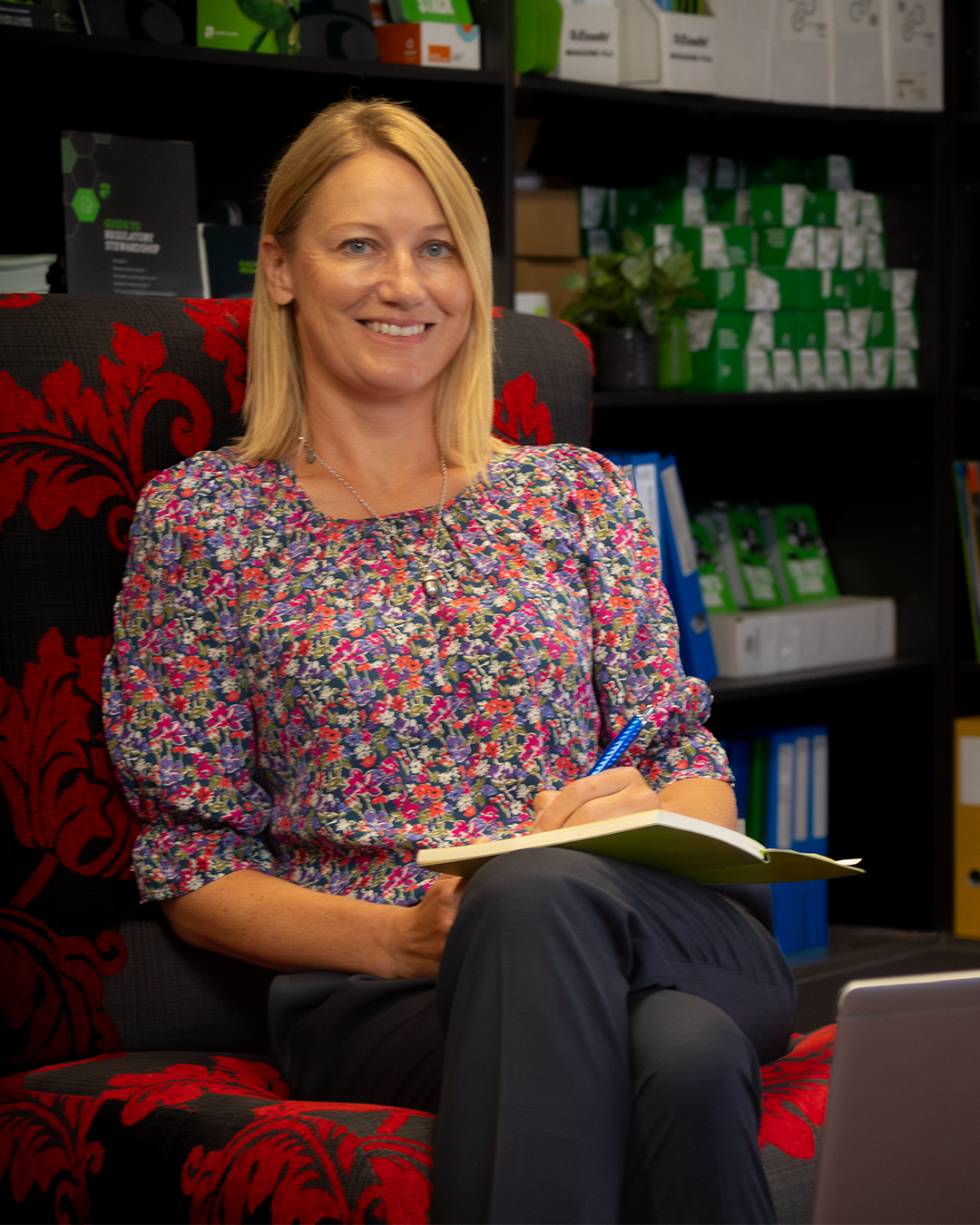 Woman in floral blouse smiling with notebook, seated in red-patterned chair with green-branded materials on shelves