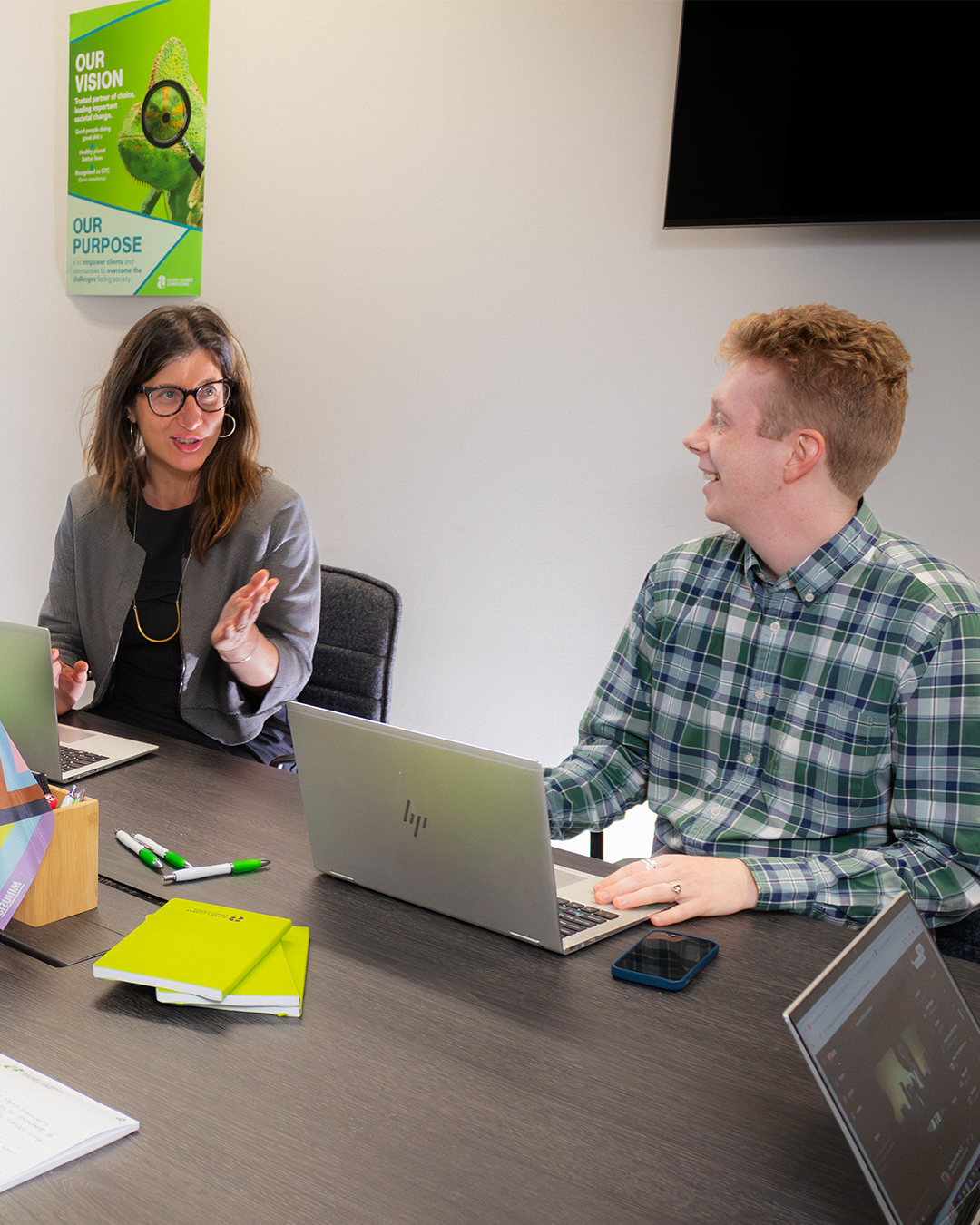 Woman in grey blazer gesturing while speaking to colleague in plaid shirt during meeting