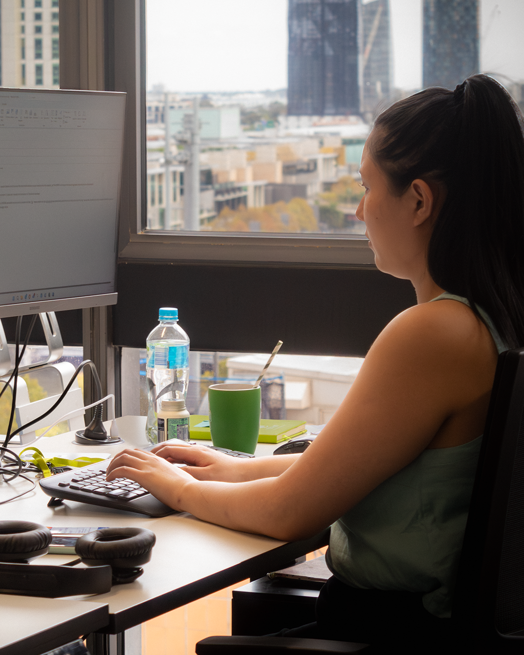 Woman looking out office window at city view, working at computer with water bottle and green mug