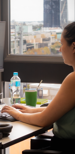Woman looking out office window at city view, working at computer with water bottle and green mug