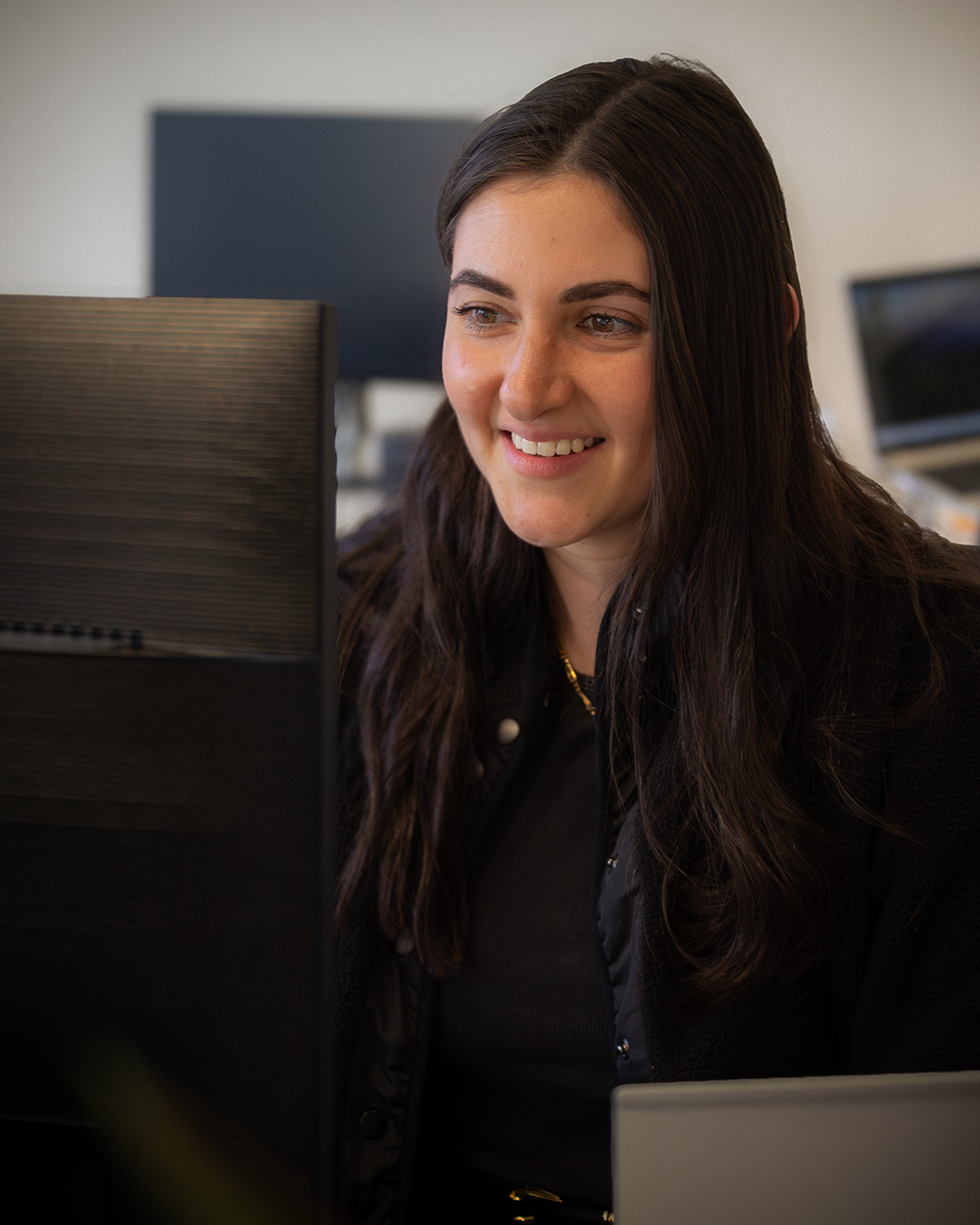 Woman with dark hair smiling at computer monitor in office setting