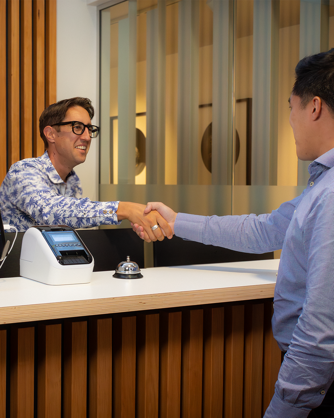 Two professionals shaking hands at reception desk, with label printer and service bell visible