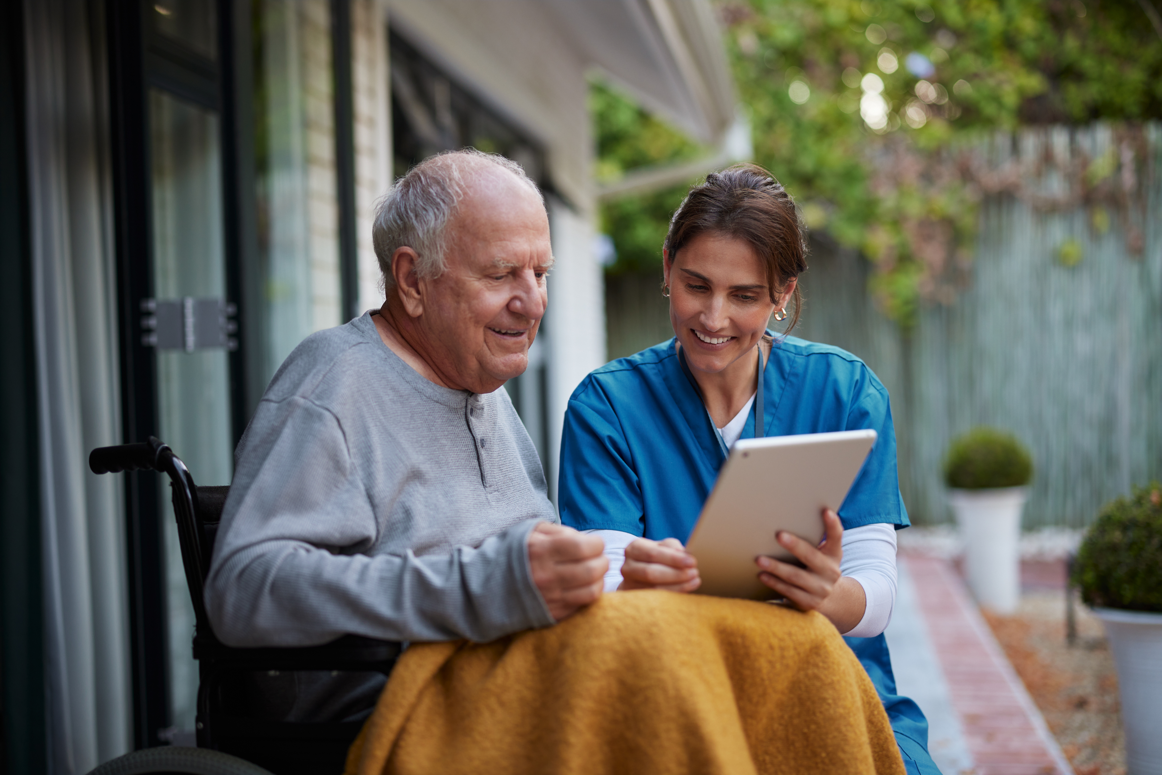 Healthcare worker and elderly man sharing a tablet moment on their porch, exemplifying supportive in-home care and digital inclusion.