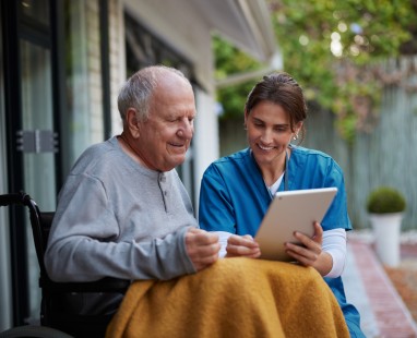 Healthcare worker and elderly man sharing a tablet moment on their porch, exemplifying supportive in-home care and digital inclusion.