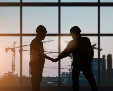 Silhouettes of two construction workers in hard hats shaking hands, with construction cranes visible at sunset behind them.
