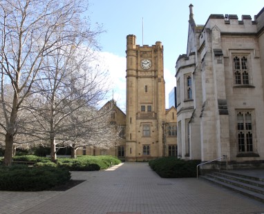 Historic stone university clocktower and buildings framing a courtyard pathway, with bare winter trees creating a scholarly atmosphere.