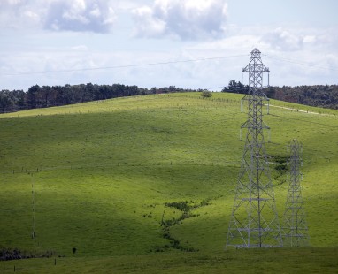 Power transmission towers standing amid rolling green farmland in rural New Zealand, with forested hills in the background and a natural gully cutting through the pasture.